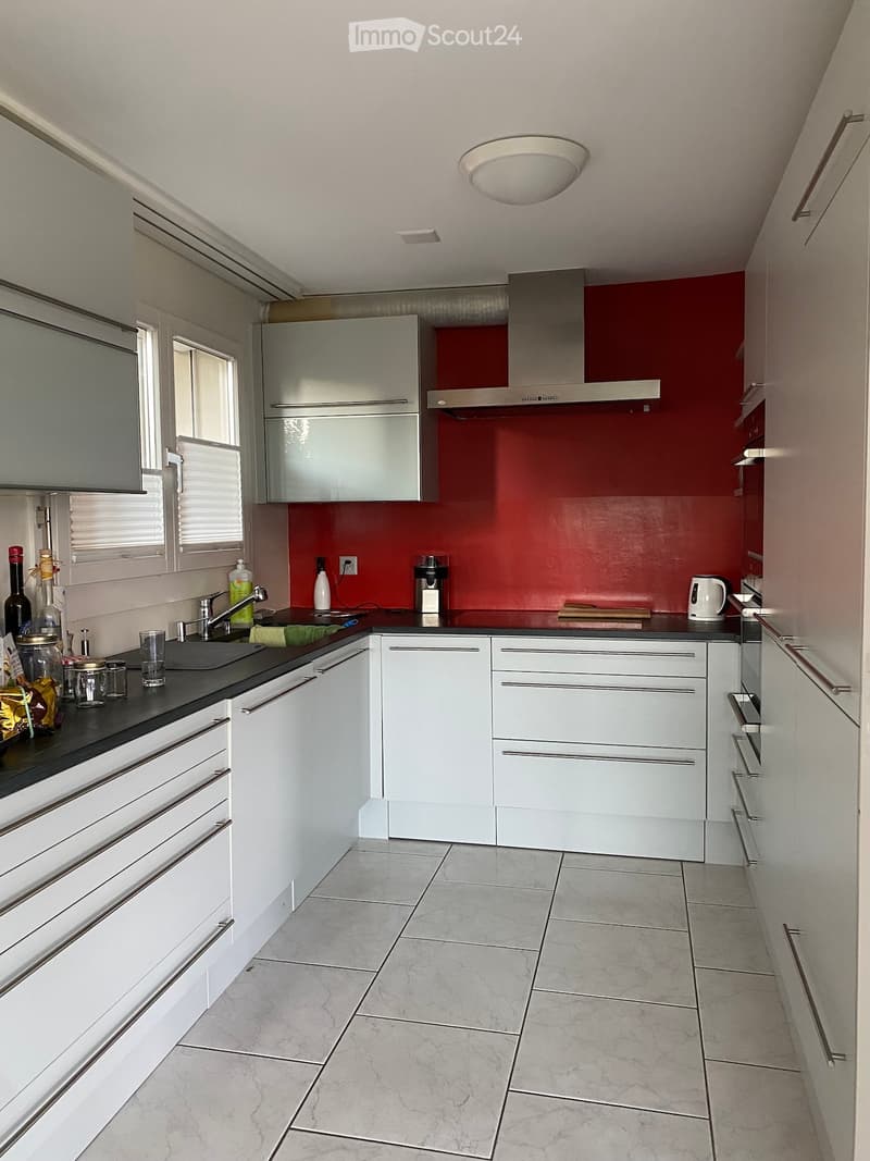 Modern kitchen with white cabinets, stainless steel appliances, and a red accent wall. Features include a sink, range hood, and various kitchen items on the counter.