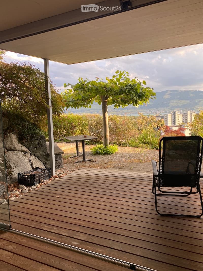 Wooden deck with a black folding chair and a small table, overlooking a scenic view of trees and mountains.