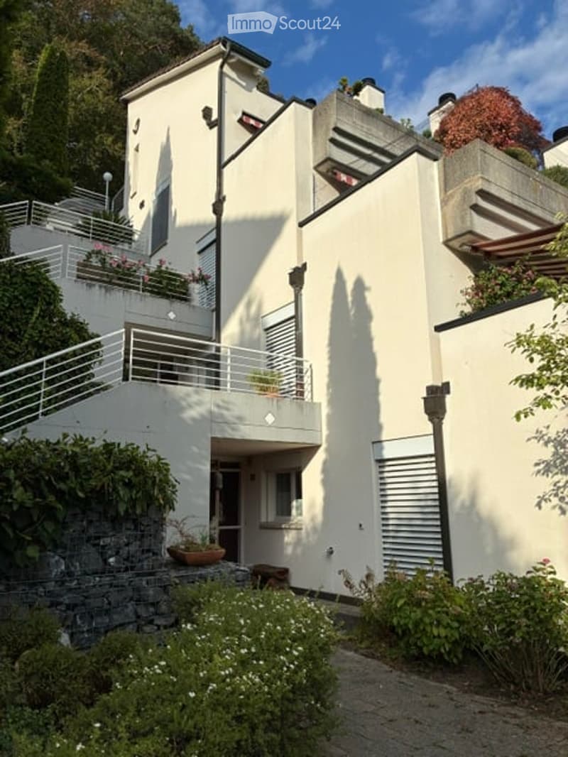 Modern two-story house with white exterior, balconies, and greenery around the entrance.