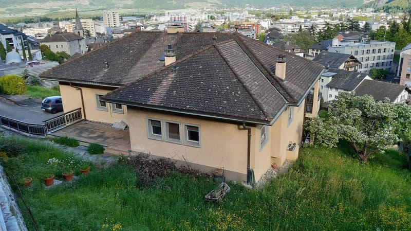 Two-story house with a tiled roof, beige exterior, multiple windows, and a small garden area with potted plants.