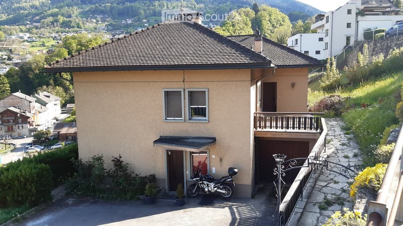 Two-story house with beige exterior, multiple windows, garage, and balcony overlooking a scenic valley with greenery and distant mountains.