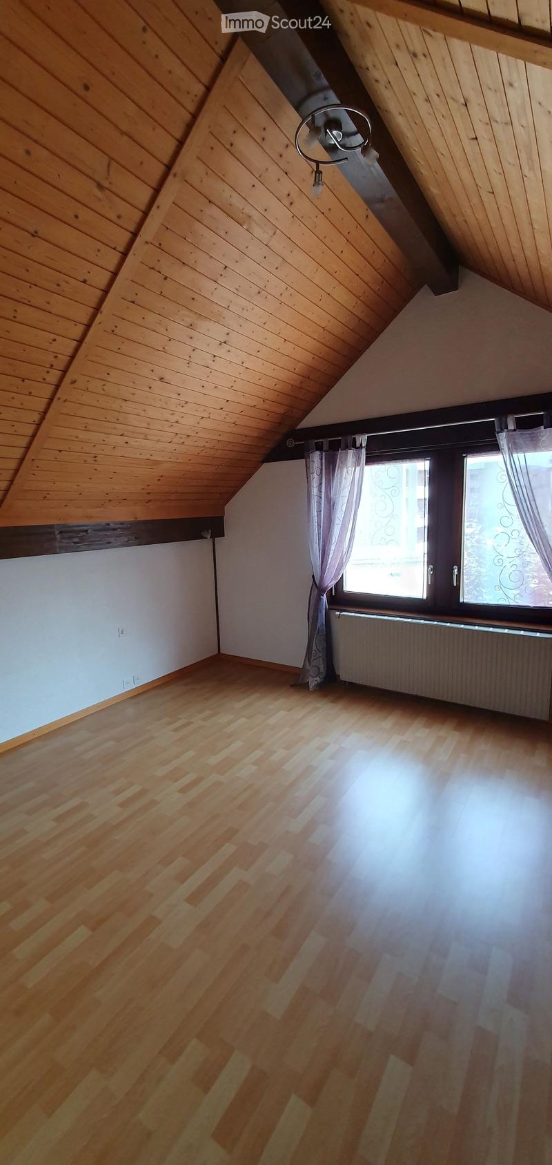 Empty attic room with wooden slatted ceiling, wooden floor, white walls, large window with curtains, radiator, and exposed wooden beams.