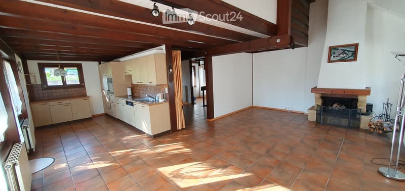 Open-plan living and dining area with a fireplace, kitchen area, and exposed wooden beams on the ceiling. The space features tiled flooring and large windows allowing natural light to fill the room.