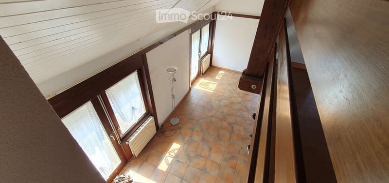 A narrow hallway with tiled flooring, wooden railings, and large windows allowing natural light to enter. The walls are painted white, and there is a floor lamp near the center of the hallway.