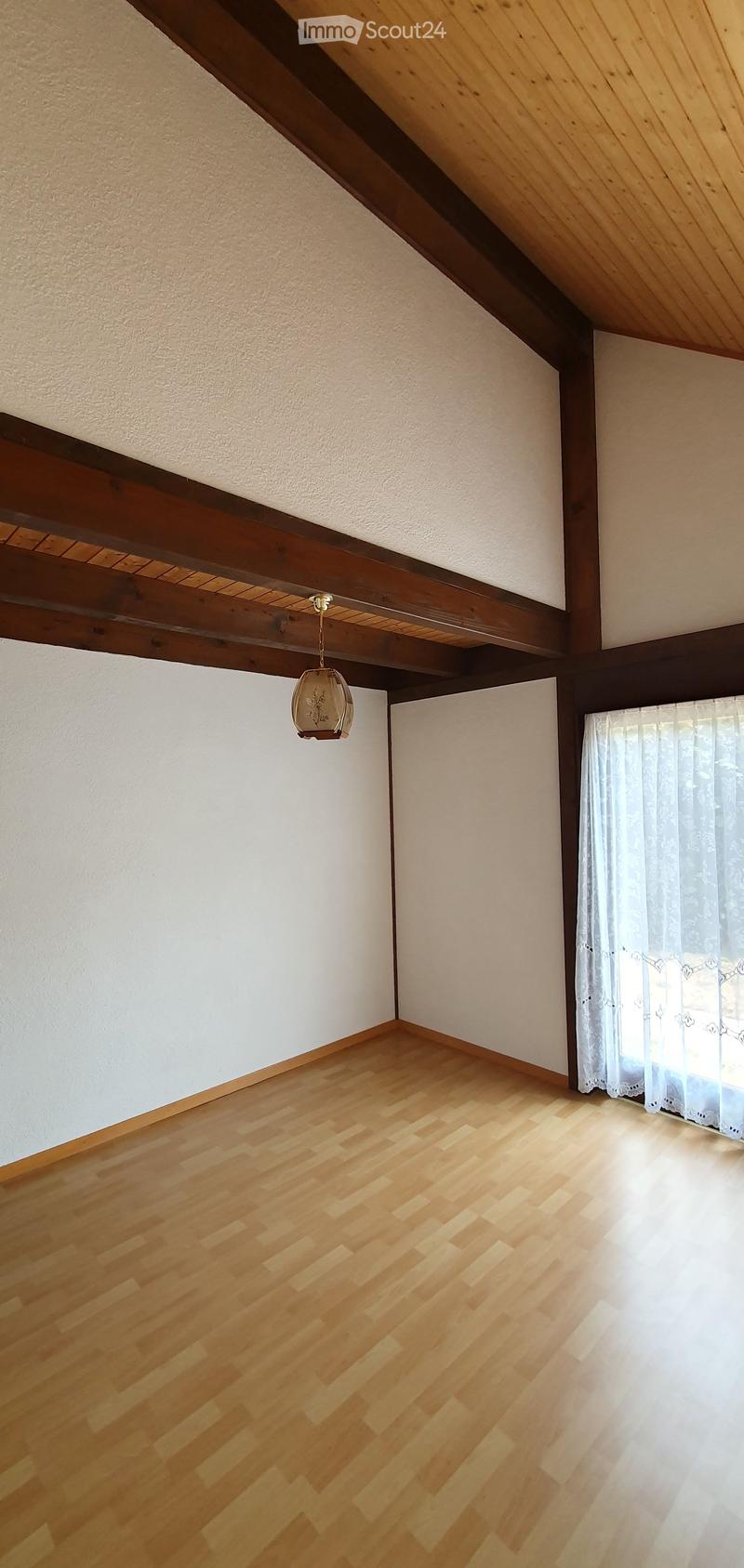 Empty room with wooden flooring, white walls, and a hanging light fixture. A window with white curtains allows natural light to enter.