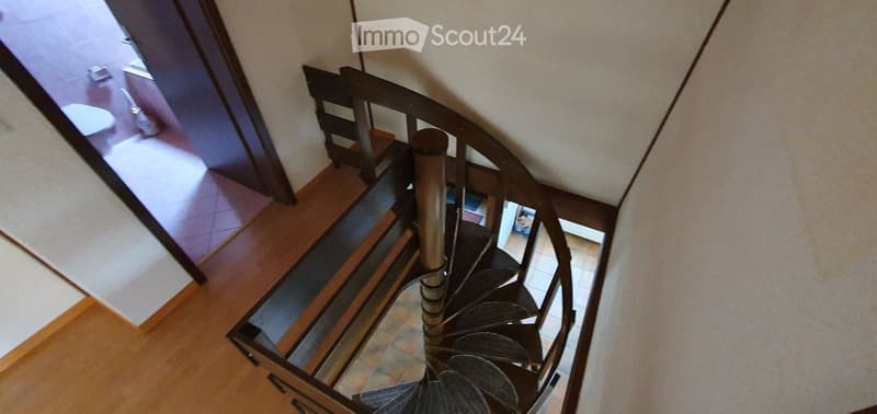 Spiral staircase with wooden steps and metal railing, leading to a bathroom visible at the top.