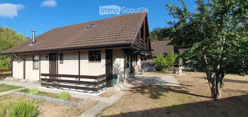 Single-story house with a tiled roof, wooden balcony, and surrounding garden area with a tree and outdoor furniture.
