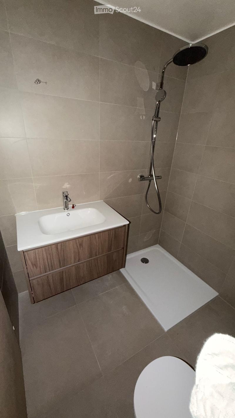 Modern bathroom with a white sink, wooden cabinet, showerhead, and toilet. Beige tiled walls and floor.