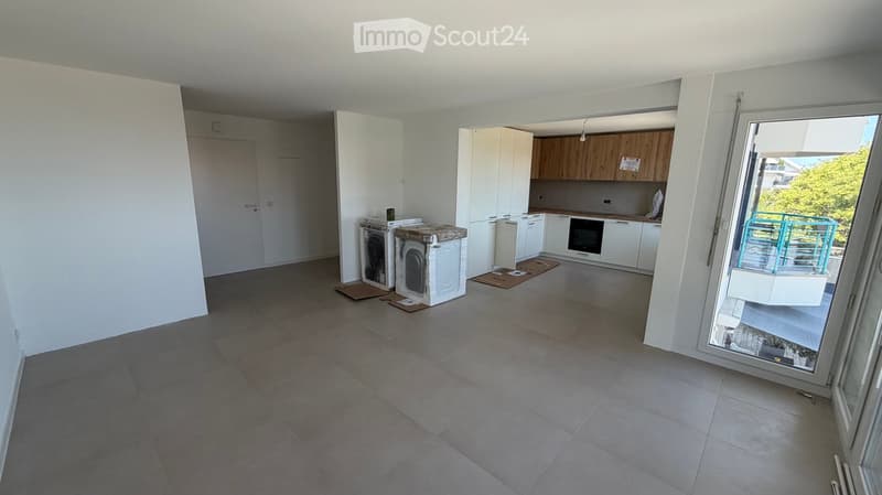 Empty room with tiled floor, white walls, and a large sliding glass door leading to a balcony. Kitchen area with cabinets and appliances is visible in the background.