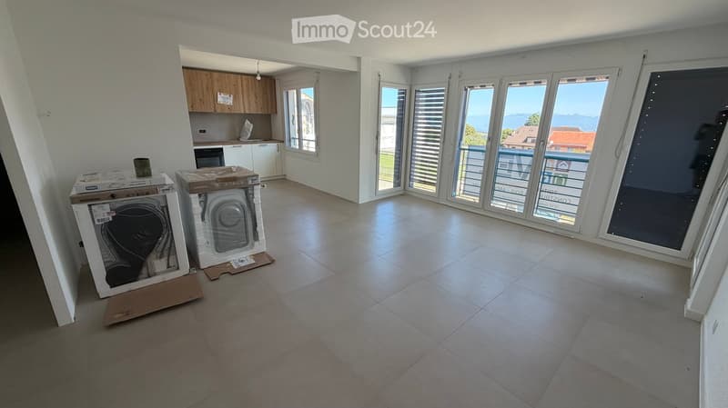 Empty room with tiled floor, large windows, and a view of the sea. Kitchen area visible in the background with cabinets and appliances.