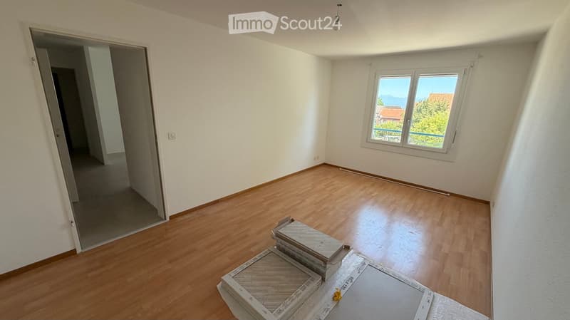 Empty room with wooden flooring, white walls, and a large window with a view of the outside. The room is unfurnished, with only a few stacked boxes and a piece of plastic sheeting on the floor.
