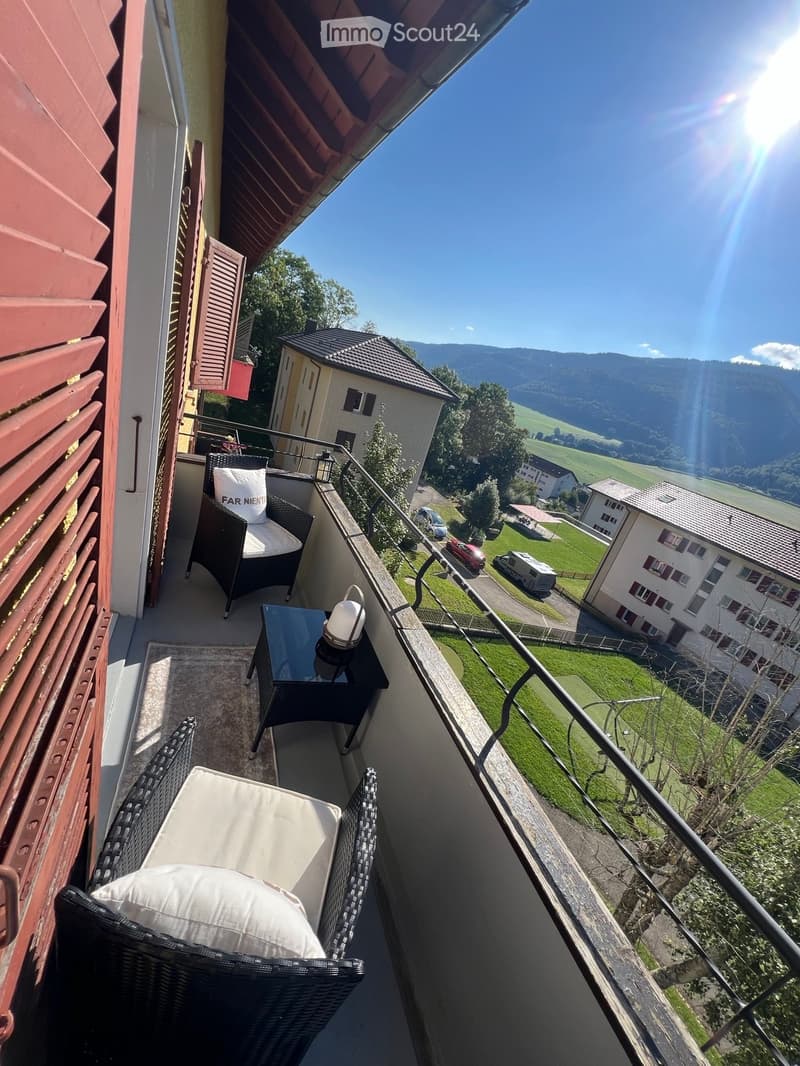 Balcony with wicker chairs, small table, and lamp, overlooking a scenic valley and mountains.