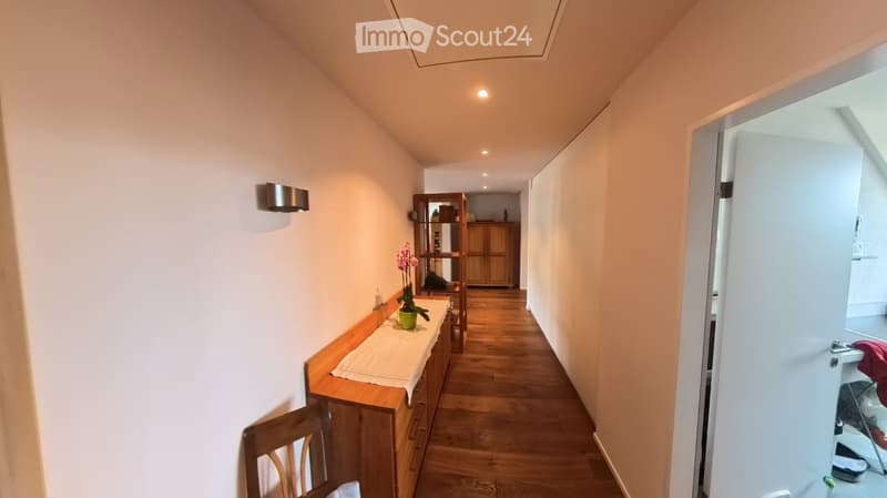 A narrow hallway with wooden flooring, light-colored walls, and built-in storage units. A small table with a vase of flowers is visible on the left side, and a partially open door leads to another room on the right.