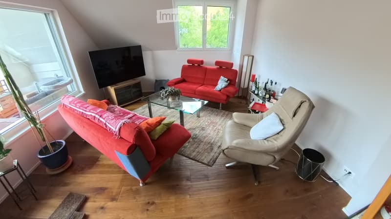 Modern living room with wooden flooring, large windows, red sofas, a glass coffee table, a leather armchair, and a television.