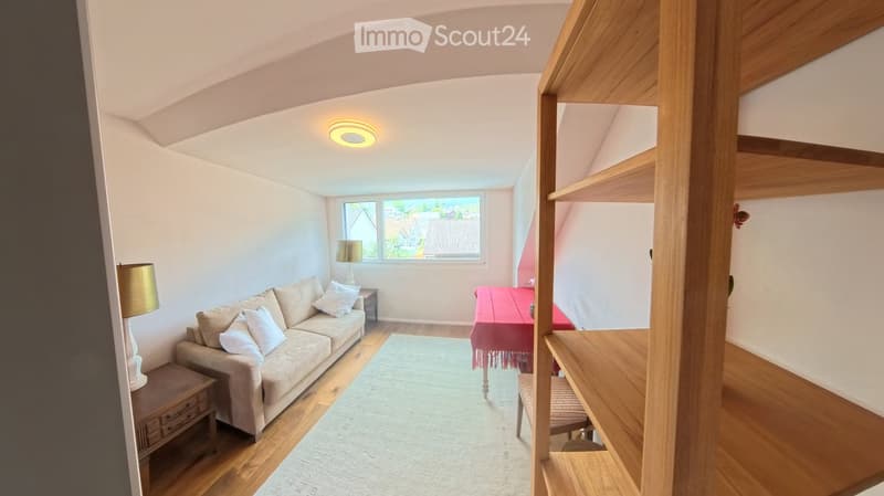 Bright living room with wooden floors, a beige sofa, a small table with a red tablecloth, and a side table with a lamp.