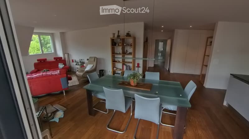 Modern living and dining area with wooden flooring, a red sofa, a glass dining table with chairs, and a bookshelf with decorative items.