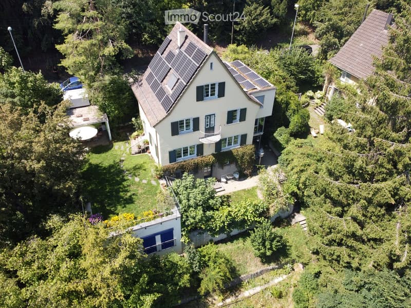 Aerial view of a two-story house with solar panels on the roof, surrounded by greenery and trees. The house features multiple windows, a balcony, and a small patio area with outdoor furniture.
