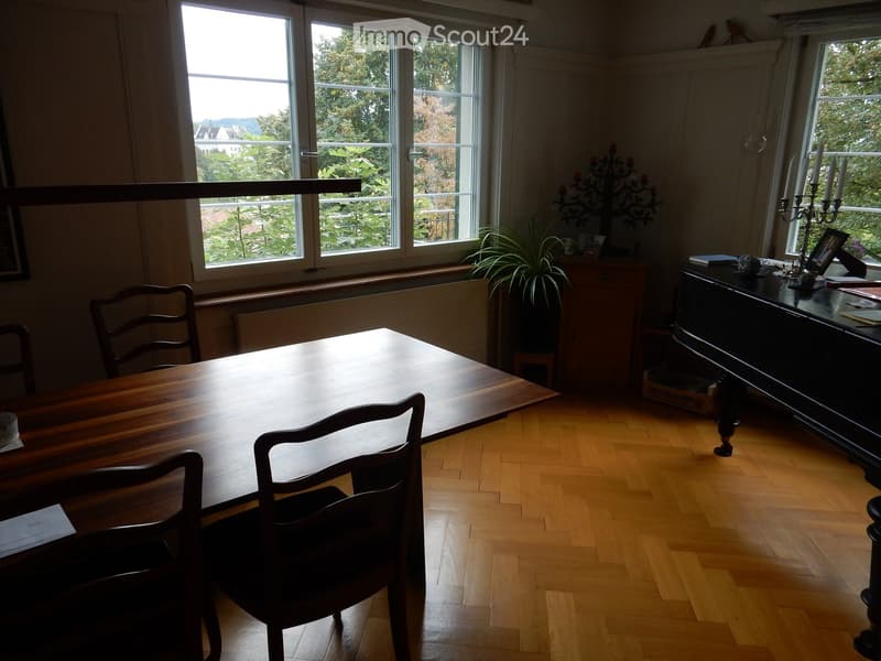 Bright room with wooden flooring, large windows offering a scenic view, a long wooden table with chairs, a piano, and decorative candle holders.