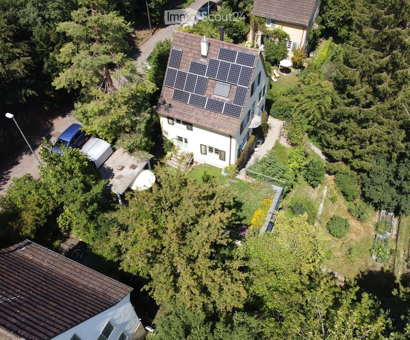 Aerial view of a two-story house with solar panels on the roof, surrounded by trees and greenery. The house has multiple windows and a small balcony. There is a driveway with parked cars and a garden area with outdoor furniture.