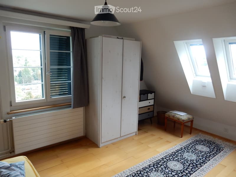 Bright bedroom with wooden flooring, large windows, a bed with pillows, a wardrobe, a small stool, a drawer unit, and a patterned rug.