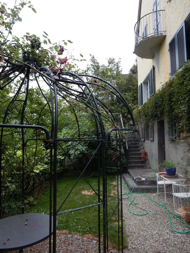 Garden area with a metal frame structure, a black table, white chairs, potted plants, and a staircase leading to a building with a balcony.