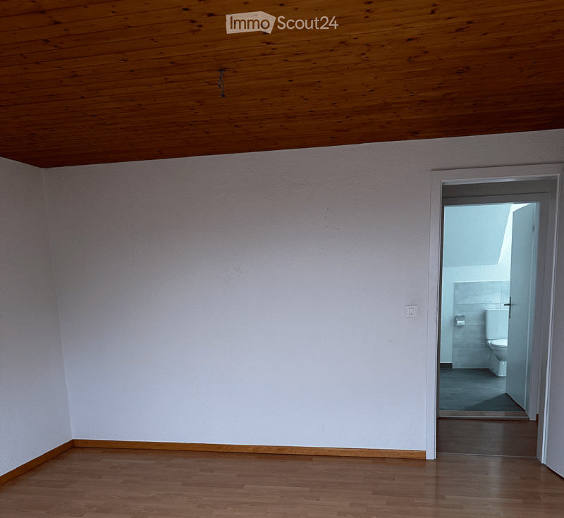 Empty room with wooden floor, white walls, wooden ceiling, door leading to a bathroom