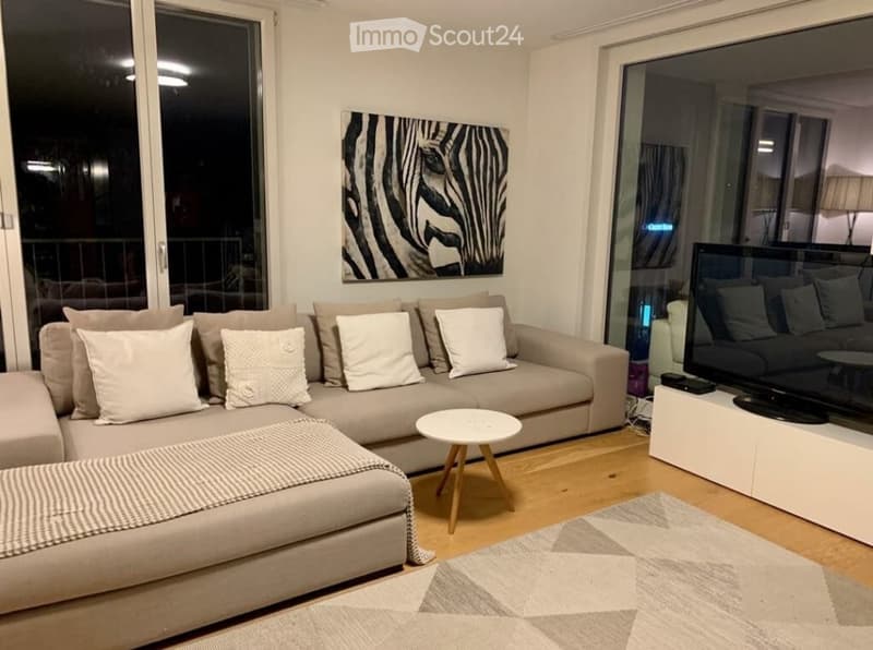 Modern living room with large windows and balcony access, featuring a beige sectional sofa, white throw pillows, a small white coffee table, a TV stand with a flat-screen TV, and a patterned rug.