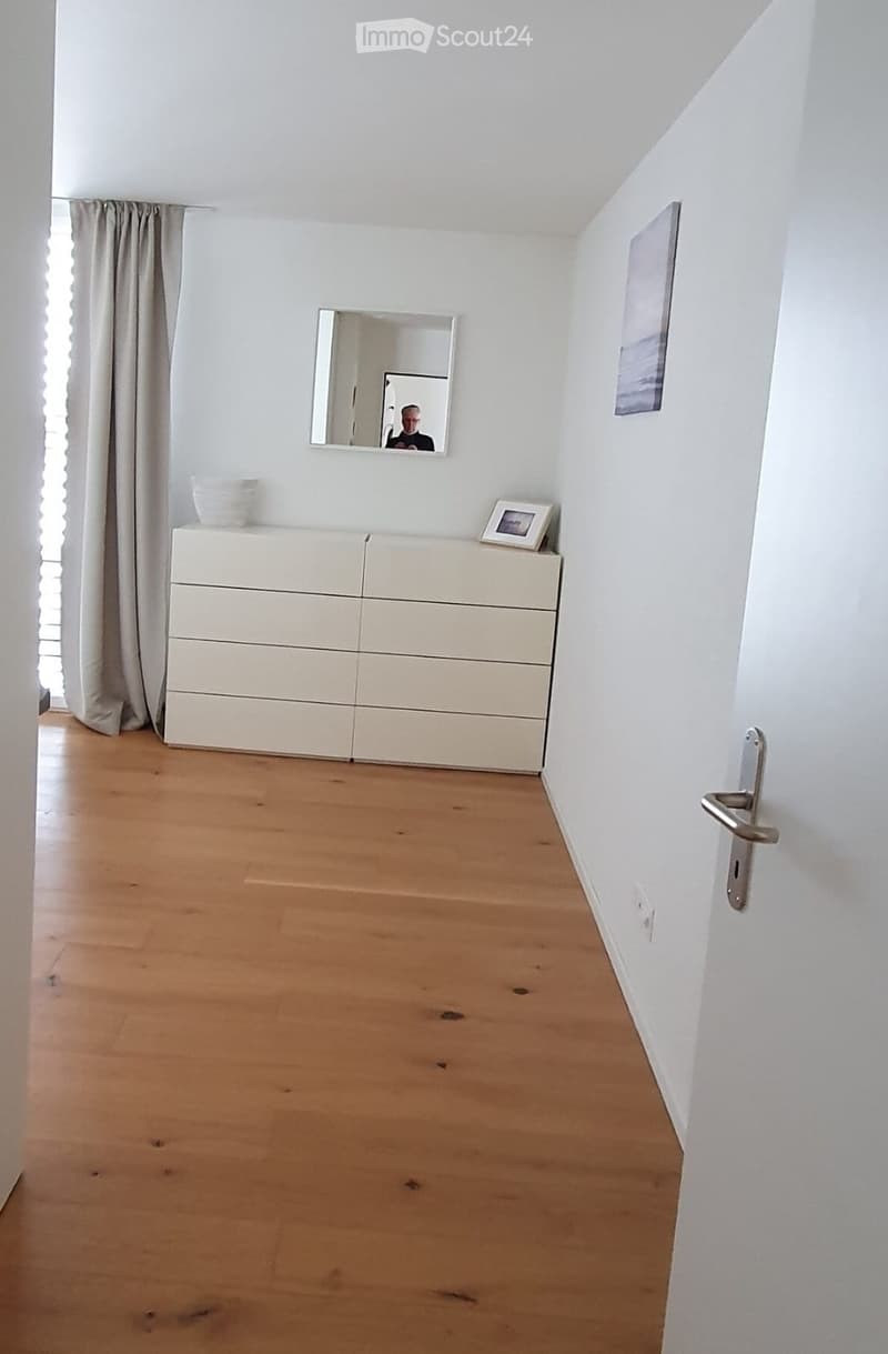 Empty room with light wooden flooring, white walls, a white dresser with drawers, a mirror, and a framed picture on the wall.