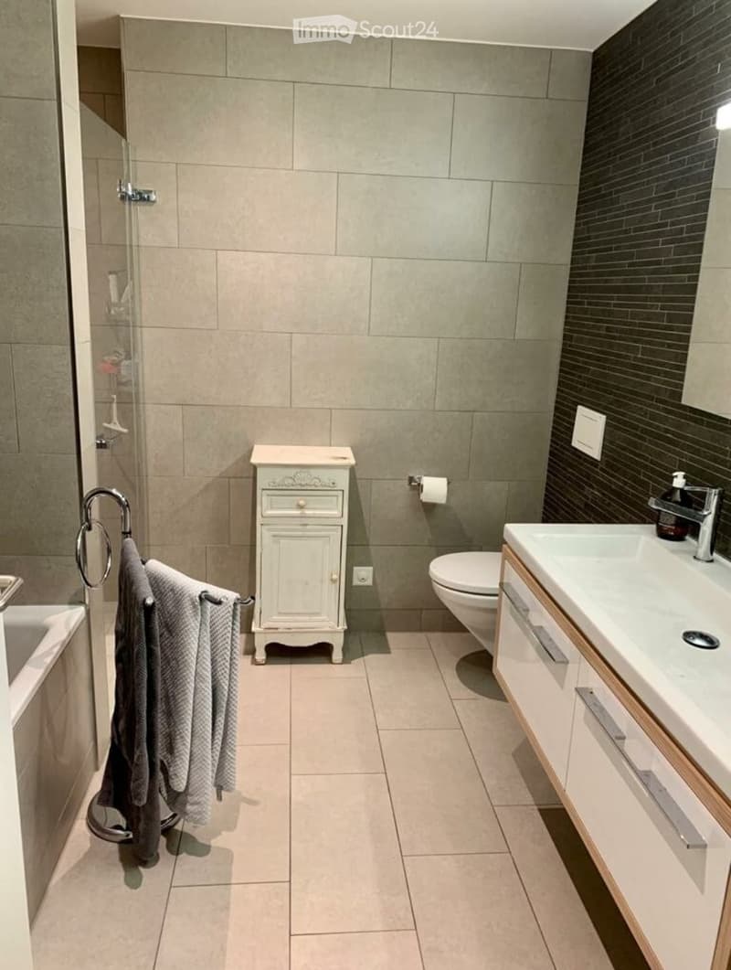 Modern bathroom with beige tiles, a white sink with wooden cabinet, a toilet, a small white cabinet, and a towel hanging on a ring.