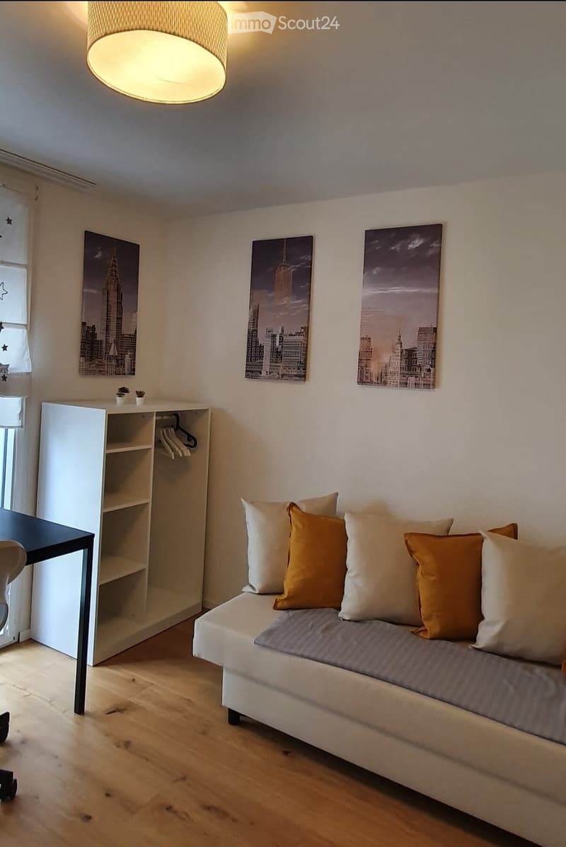 A modern living room with a white sofa, decorative pillows, a small table, a chair, a shelf with hanging clothes, and several cityscape picture frames on the wall.