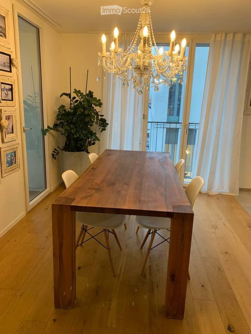 Dining room with a wooden table, white chairs, chandelier, and large windows leading to a balcony.