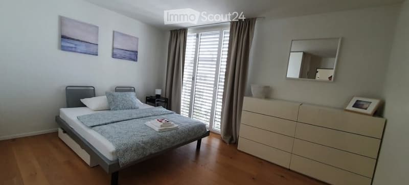 Cozy bedroom with a double bed, nightstand, dresser, mirror, and artwork on the walls. Large windows with blinds and curtains provide natural light.