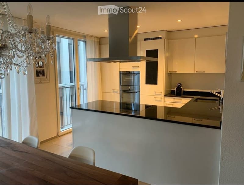 modern kitchen with glass top counter, white cabinets, black granite counter top, silver range hood, two built-in ovens, chandelier and glass door leading to a balcony