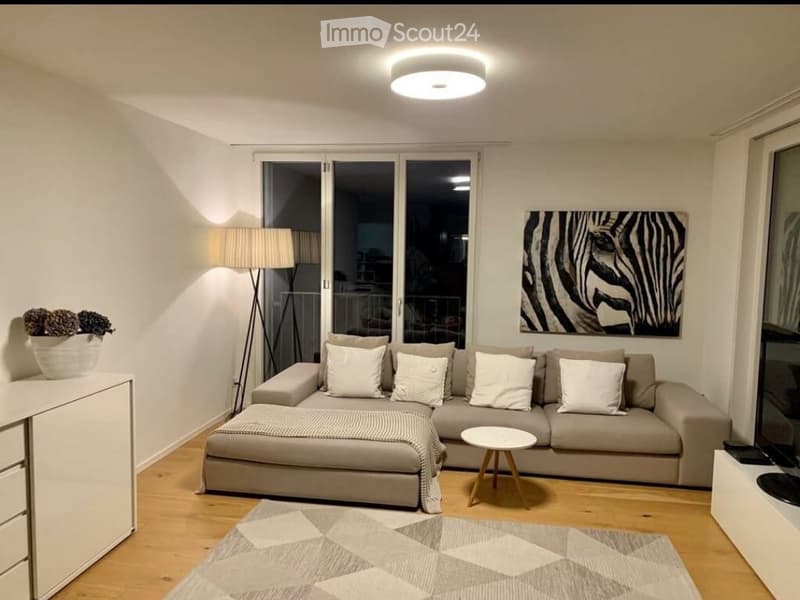 Modern, cozy living room with a sofa, a table, a lamp, and a carpet. The living room has a balcony and offers a view of the outdoors.