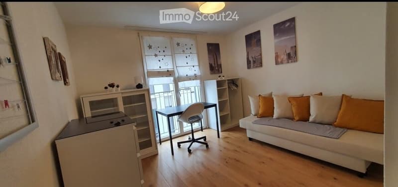 small living room with couch, white shelves, desk and chair, wooden floor, windows with curtains, paintings