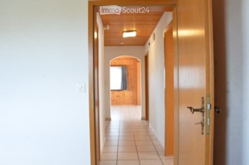 Empty hallway with wooden doors, tiled floor, and recessed lighting. The hallway leads to another room with visible furniture.