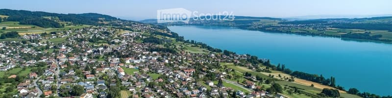 Aerial view of a town with numerous houses, surrounded by green fields and a large lake, with distant mountains visible.