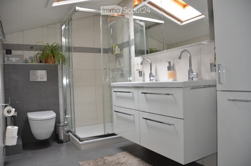 Modern bathroom with a white vanity, double sink, glass-enclosed shower, toilet, and a potted plant on the windowsill. The room features grey tiles and sleek, contemporary fixtures.