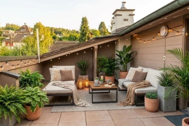 Cozy outdoor patio with two lounge chairs, coffee table, throw blankets, potted plants, string lights, and a view of trees and neighboring houses.