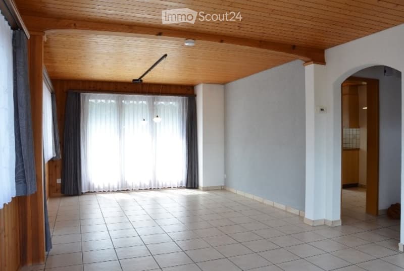 Empty room with wooden ceiling, tiled floor, large window with curtains, and an archway leading to another room.