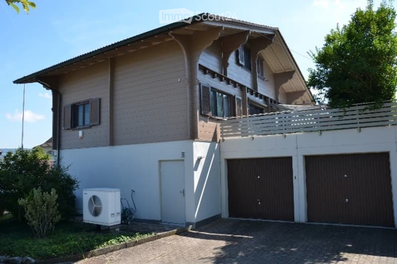 Two-story house with beige siding, white trim, and two garage doors. Features a balcony on the upper level and an air conditioning unit near the entrance.