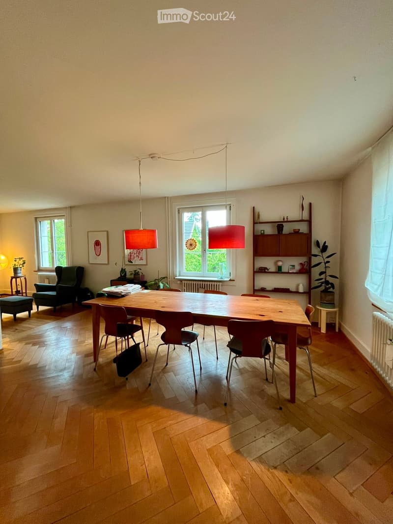 Bright dining room with wooden floor, large wooden dining table surrounded by chairs, sofa, small stool, shelves with decorative items, and plants near the windows.
