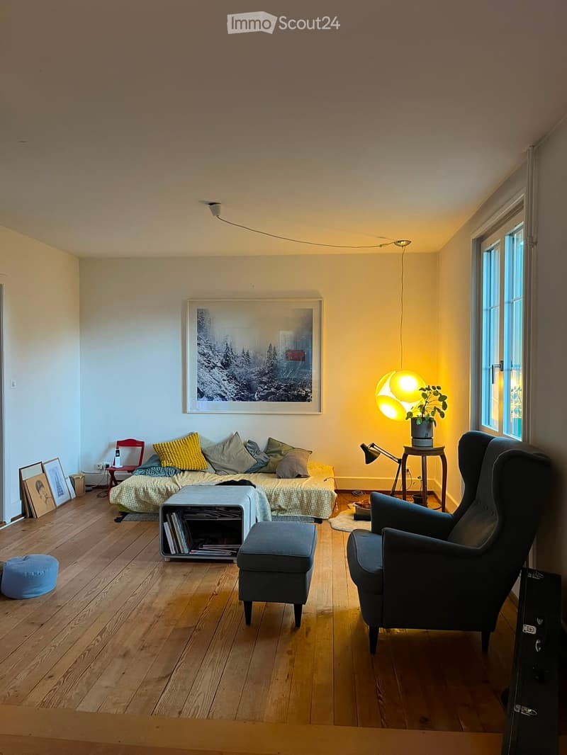 Cozy living room with wooden flooring, a sofa with pillows, a blue ottoman, a dark armchair, a small table with a lamp, and a bookshelf.