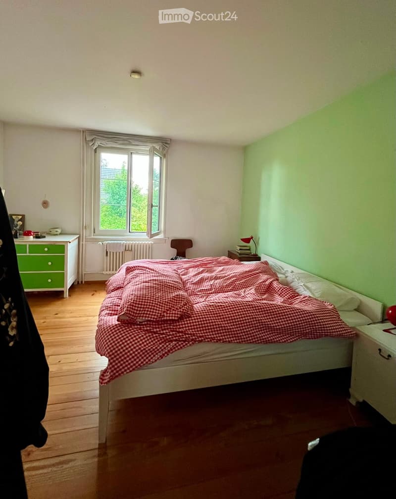 Bedroom with wooden floor, large window, bed with red checkered blanket, nightstand with lamp, small table with books, and a green drawer unit.