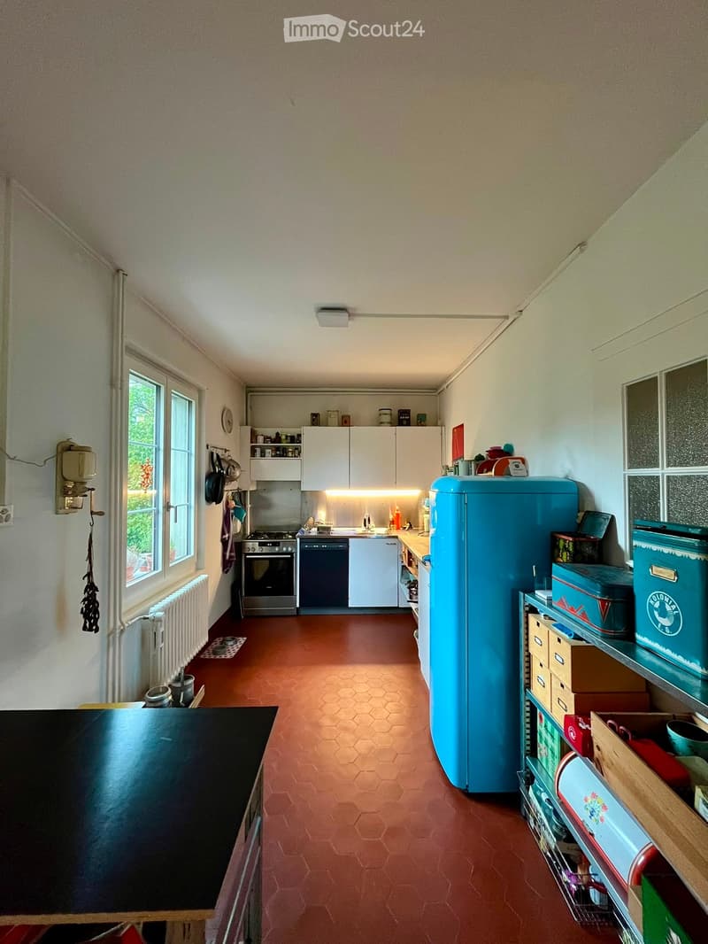 A well-lit kitchen with white cabinets, a blue refrigerator, and a black table. The floor is red hexagonal tiles, and there are shelves with various items.