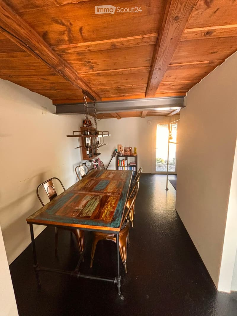 A modern dining area with a wooden table, metal chairs, and a shelf with books and decorative items. The room features exposed wooden beams on the ceiling and a large window letting in natural light.