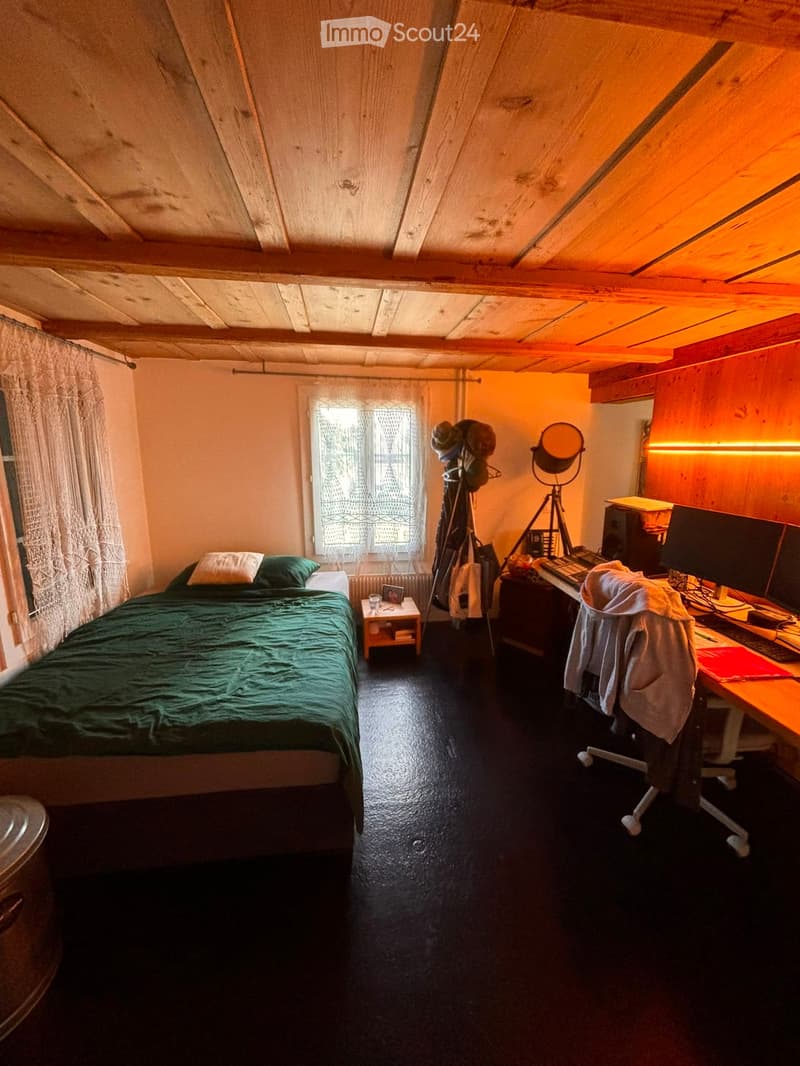 Cozy bedroom with wooden slanted ceilings, a bed with green bedding, a small nightstand, a desk with a computer setup, and a tripod with a mirror nearby.