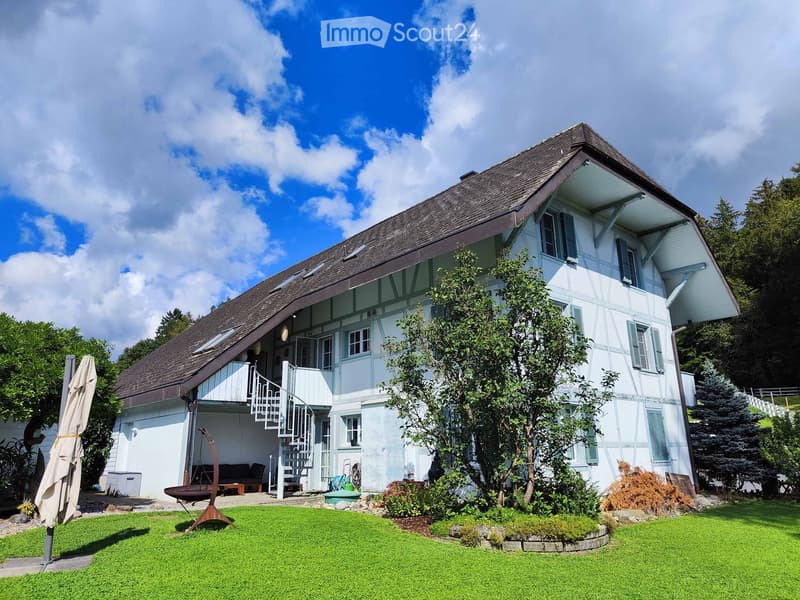 Two-story white house with a dark roof, multiple windows, and a balcony accessible via an outdoor staircase. The house is surrounded by a well-maintained lawn, trees, and shrubs.
