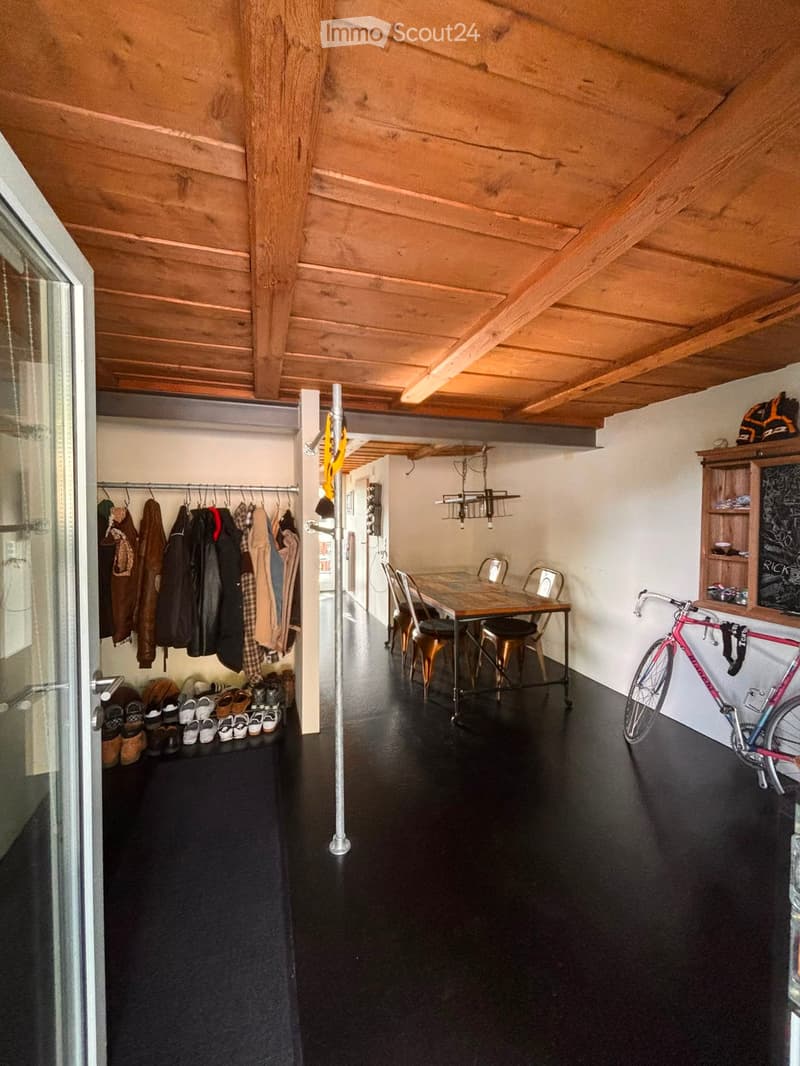 A modern, open-plan space with a wooden table and chairs, a bicycle, a coat rack, and a shelf with items. The ceiling has exposed wooden beams, and the floor is dark.