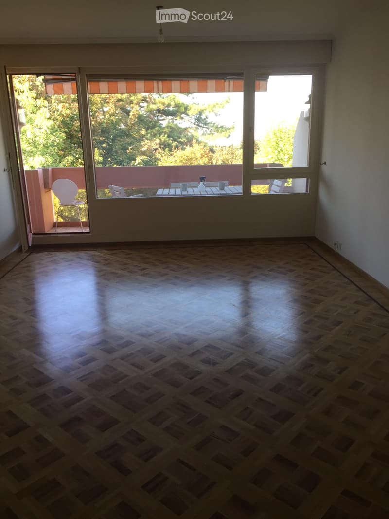 Empty room with wooden flooring, large windows, and an open door leading to a balcony with outdoor furniture.